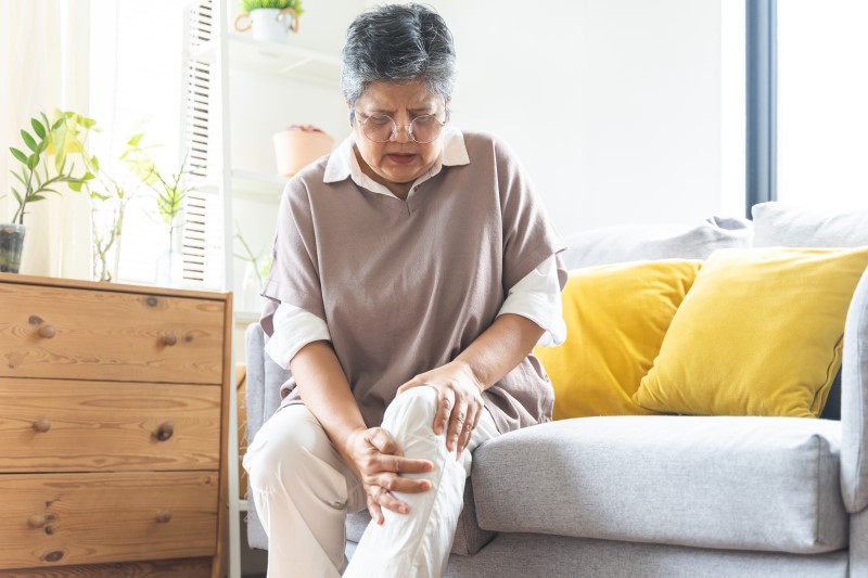 Osoba siedząca na kanapie, trzymająca się za kolano, wyrażająca ból lub dyskomfort.