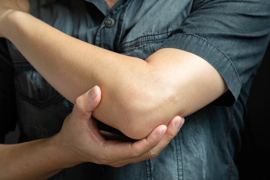 Na zdjęciu widać osobę dotykającą swojego łokcia, co może sugerować dolegliwości związane z zapaleniem stawu łokciowego. Dłoń wspiera łokieć, co często jest gestem towarzyszącym dyskomfortowi lub bólowi w tym obszarze.